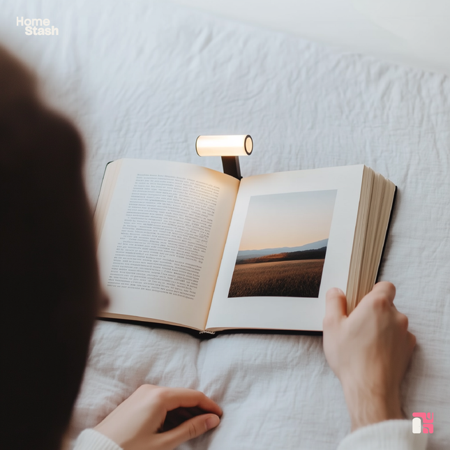 Rechargeable book light clipped to an open book, illuminating the pages. The book is resting on a white bedspread, and soft lighting creates a cozy reading atmosphere.