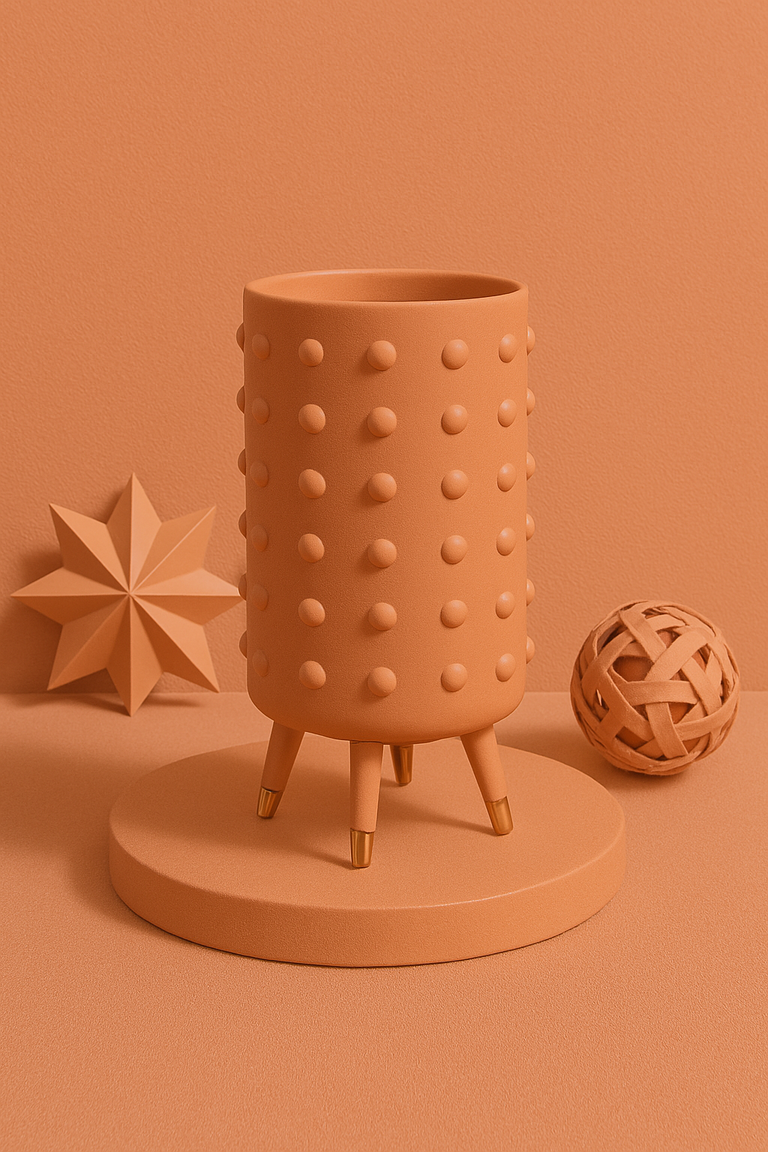 Contemporary decor scene featuring a textured orange planter with gold-tipped legs, flanked by a geometric star sculpture and woven orb on a monochrome background.
