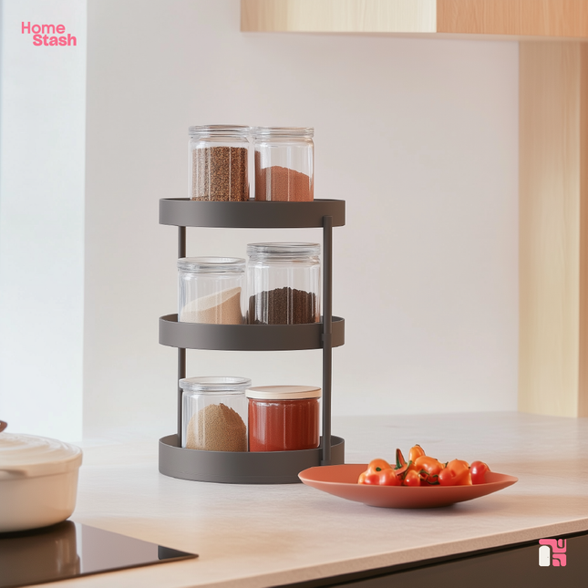 Rotating spice rack with three tiers holding glass jars filled with spices. The rack is placed on a modern kitchen countertop near a plate of fresh tomatoes. Minimalist design with warm natural lighting and clean kitchen decor.