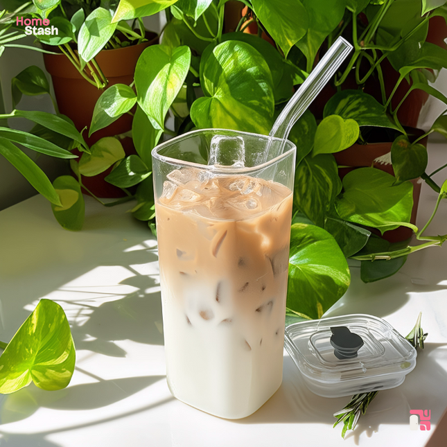 Square transparent coffee cup with iced latte and glass straw, shown beside a secure lid in natural sunlight with green potted plants in the background.
