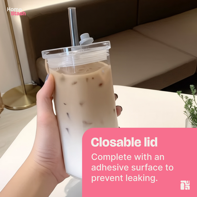 Hand holding a square transparent coffee cup filled with iced latte, featuring a closable leakproof lid and glass straw, styled in a cozy indoor setting.